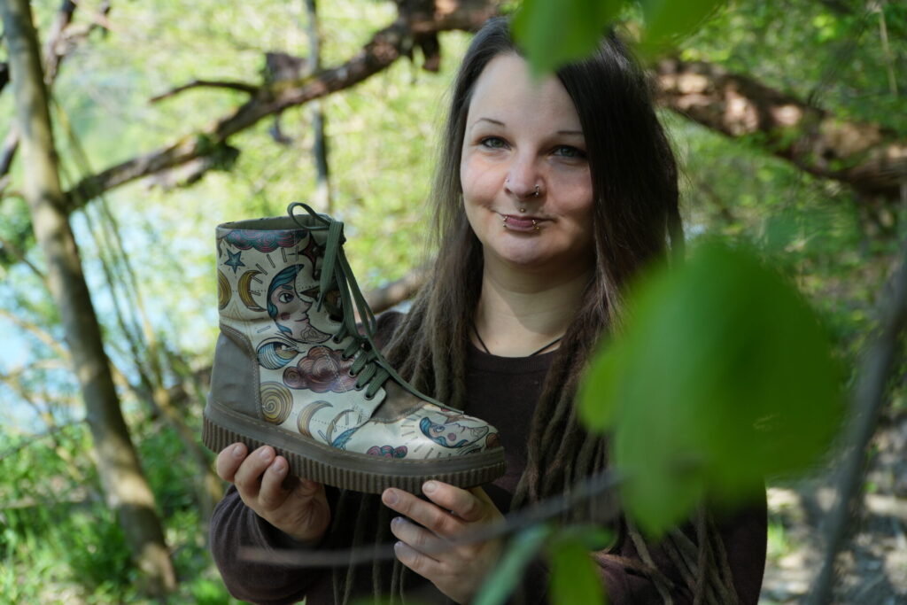 hippie mama mit piercings und dreads in erdigen farben hält bunten veganen dogo schuhe fest in der natur 