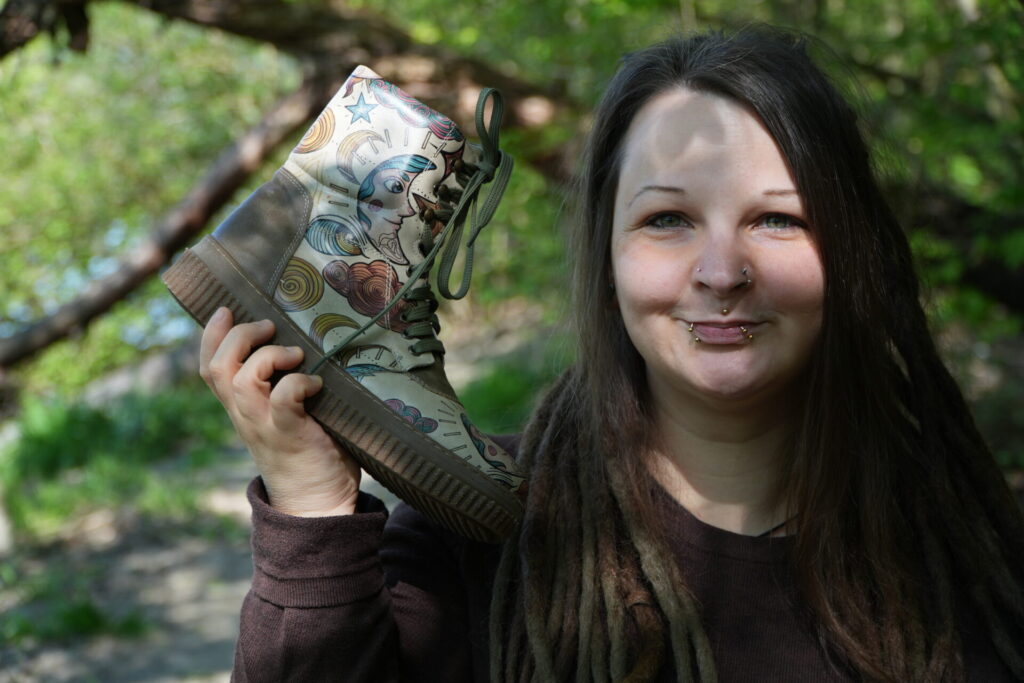hippie mama mit piercings und dreads in erdigen farben hält bunten veganen dogo schuhe fest in der natur 