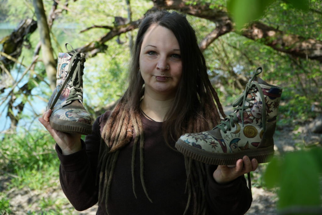 hippie mama mit piercings und dreads in erdigen farben hält bunten veganen dogo schuhe fest in der natur 
